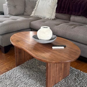 Pode incluir: Uma mesa de centro em forma de rim feita de madeira castanha escura com uma superf&iacute;cie lisa e duas pernas verticais em painel. Sobre a mesa encontra-se um vaso de cer&acirc;mica branca texturada dentro de uma ta&ccedil;a cinzenta rasa e dois comandos.