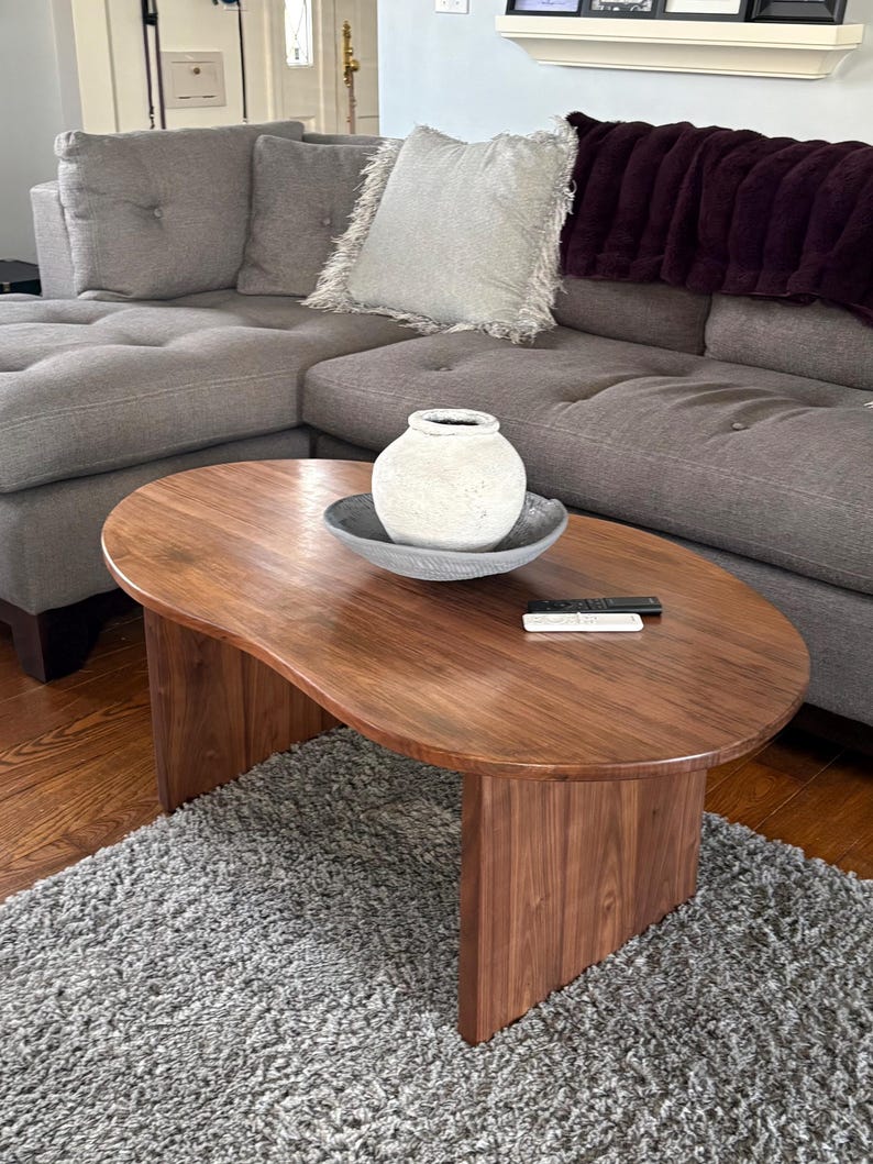 Pode incluir: Uma mesa de centro em forma de rim feita de madeira castanha escura com uma superf&iacute;cie lisa e duas pernas verticais em painel. Sobre a mesa encontra-se um vaso de cer&acirc;mica branca texturada dentro de uma ta&ccedil;a cinzenta rasa e dois comandos.
