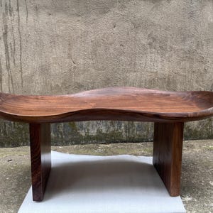 May include: A low wooden bench crafted from dark brown walnut wood, featuring a smooth, undulating, wave-like seat surface with a natural grain pattern. The bench is supported by two solid, vertical rectangular legs on each side.