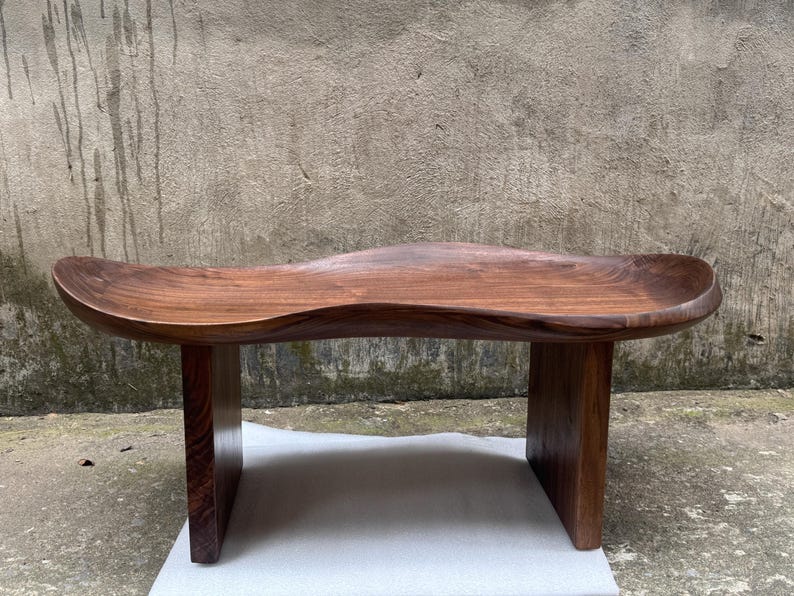 May include: A low wooden bench crafted from dark brown walnut wood, featuring a smooth, undulating, wave-like seat surface with a natural grain pattern. The bench is supported by two solid, vertical rectangular legs on each side.