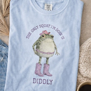 Puede incluir: Una camiseta azul claro con un gr&aacute;fico de una rana verde que lleva un sombrero de vaquero rosa, un cintur&oacute;n rosa y botas de vaquero rosas. El texto impreso en morado alrededor de la rana dice, The only squat I'm doin' is diddly. La camiseta tiene cuello redondo.