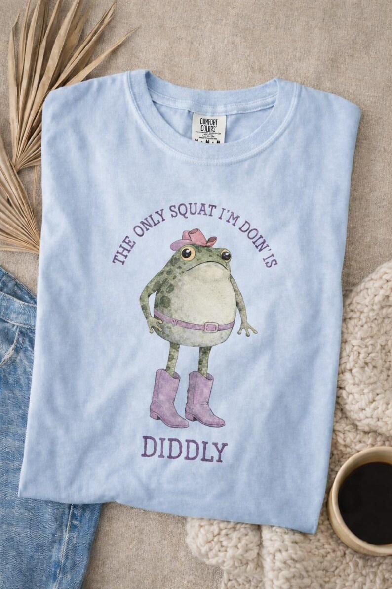 Puede incluir: Una camiseta azul claro con un gr&aacute;fico de una rana verde que lleva un sombrero de vaquero rosa, un cintur&oacute;n rosa y botas de vaquero rosas. El texto impreso en morado alrededor de la rana dice, The only squat I'm doin' is diddly. La camiseta tiene cuello redondo.