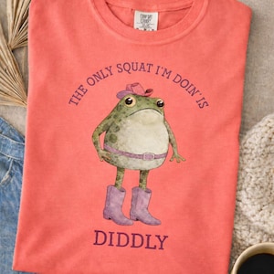 Puede incluir: Una camiseta de manga corta color coral con el gr&aacute;fico de una rana verde que lleva un sombrero de vaquero rosa, un cintur&oacute;n rosa y botas de vaquero moradas. El texto sobre la rana dice THE ONLY SQUAT I'M DOIN' IS y debajo DIDDLY.
