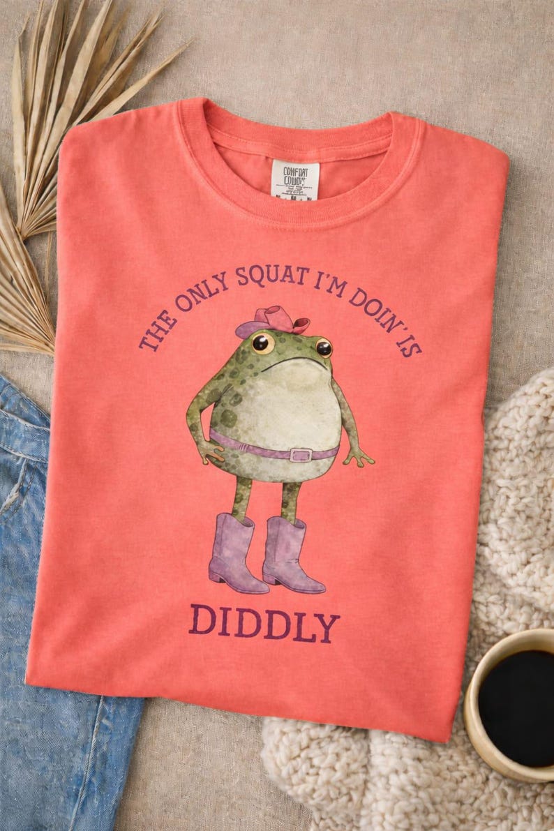 Puede incluir: Una camiseta de manga corta color coral con el gr&aacute;fico de una rana verde que lleva un sombrero de vaquero rosa, un cintur&oacute;n rosa y botas de vaquero moradas. El texto sobre la rana dice THE ONLY SQUAT I'M DOIN' IS y debajo DIDDLY.
