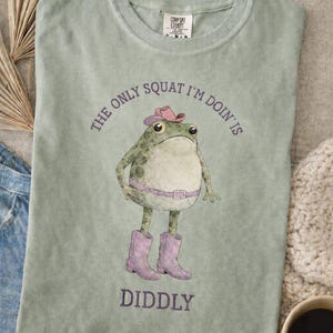 Puede incluir: Una camiseta verde salvia con el gr&aacute;fico de una rana que lleva un sombrero de vaquero rosa, un cintur&oacute;n rosa y botas de vaquero rosas. El texto sobre la rana dice THE ONLY SQUAT I'M DOIN' IS y debajo dice DIDDLY en tinta morada oscura.