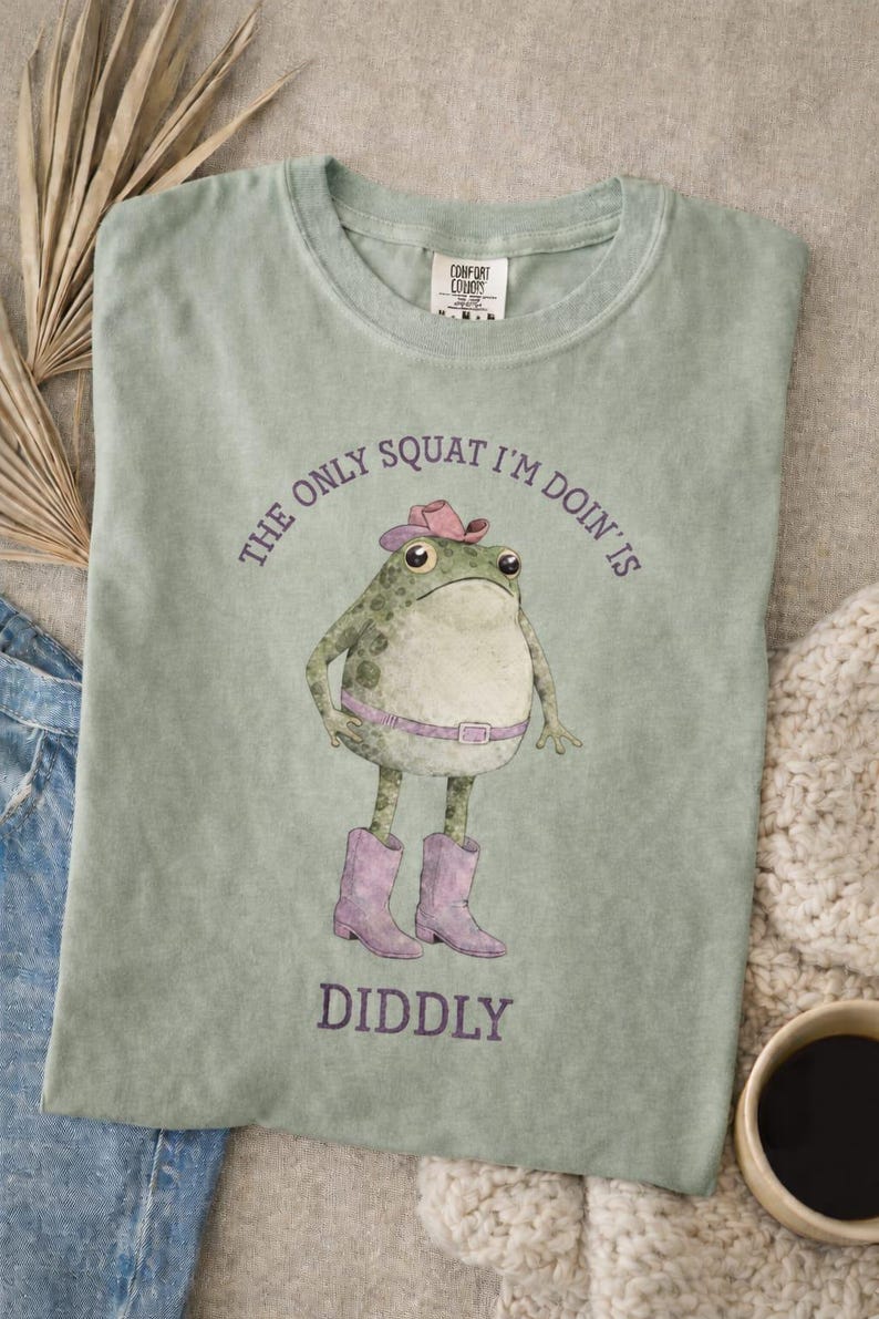 Puede incluir: Una camiseta verde salvia con el gr&aacute;fico de una rana que lleva un sombrero de vaquero rosa, un cintur&oacute;n rosa y botas de vaquero rosas. El texto sobre la rana dice THE ONLY SQUAT I'M DOIN' IS y debajo dice DIDDLY en tinta morada oscura.