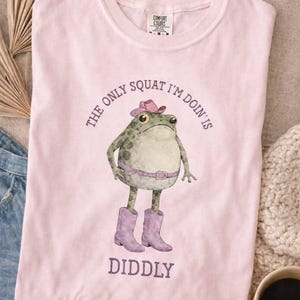 Puede incluir: Una camiseta de manga corta de color rosa claro con un gr&aacute;fico de una rana verde que lleva un sombrero de vaquero rosa, un cintur&oacute;n rosa y botas de vaquero moradas. El texto sobre la rana dice THE ONLY SQUAT I'M DOIN' IS y debajo DIDDLY.