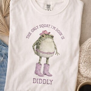 Puede incluir: Una camiseta blanca con un gr&aacute;fico de una rana verde que lleva un sombrero de vaquero rosa, un cintur&oacute;n rosa y botas de vaquero rosas. El texto sobre la rana dice THE ONLY SQUAT I'M DOIN' IS y el texto debajo dice DIDDLY en letras moradas.