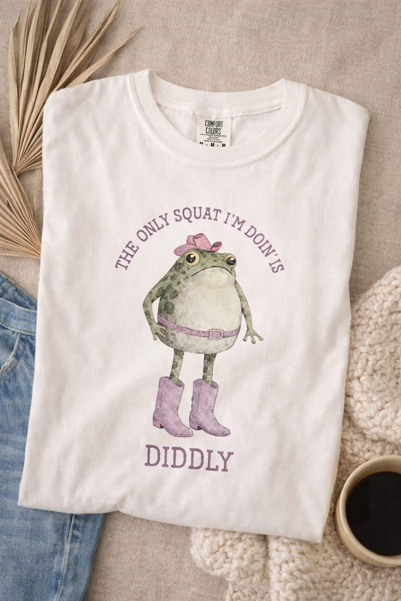 Puede incluir: Una camiseta blanca con un gr&aacute;fico de una rana verde que lleva un sombrero de vaquero rosa, un cintur&oacute;n rosa y botas de vaquero rosas. El texto sobre la rana dice THE ONLY SQUAT I'M DOIN' IS y el texto debajo dice DIDDLY en letras moradas.