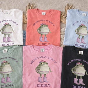 Puede incluir: Seis camisetas de cuello redondo en blanco, coral, azul claro, verde salvia, rosa y negro. Cada camiseta presenta un gr&aacute;fico de una rana con sombrero y botas rosas con el texto, THE ONLY SQUAT I'M DOIN' IS DIDDLY, impreso en color morado.
