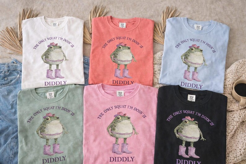 Puede incluir: Seis camisetas de cuello redondo en blanco, coral, azul claro, verde salvia, rosa y negro. Cada camiseta presenta un gr&aacute;fico de una rana con sombrero y botas rosas con el texto, THE ONLY SQUAT I'M DOIN' IS DIDDLY, impreso en color morado.