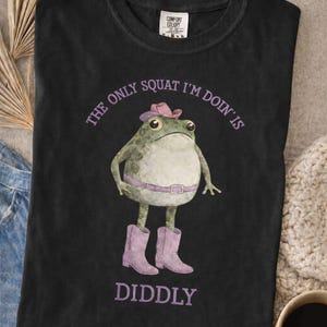 Puede incluir: Una camiseta negra con el gr&aacute;fico de una rana verde que lleva un sombrero de vaquero rosa, un cintur&oacute;n rosa y botas de vaquero rosas. El texto sobre la rana dice THE ONLY SQUAT I'M DOIN'S y debajo de la rana dice DIDDLY en letras moradas.