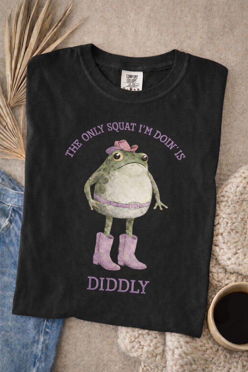 Puede incluir: Una camiseta negra con el gr&aacute;fico de una rana verde que lleva un sombrero de vaquero rosa, un cintur&oacute;n rosa y botas de vaquero rosas. El texto sobre la rana dice THE ONLY SQUAT I'M DOIN'S y debajo de la rana dice DIDDLY en letras moradas.