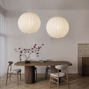 Puede incluir: Dos grandes lámparas colgantes redondas con una pantalla acanalada de color crema cuelgan sobre una mesa de comedor de madera. La mesa está rodeada de sillas de color claro. Un jarrón con flores rosas se encuentra sobre la mesa, agregando un toque de color a la habitación de tonos neutros.