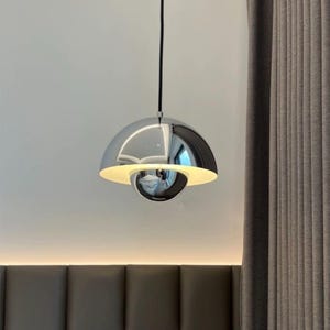 May include: A contemporary pendant light with a chrome finish. The light features a reflective, domed top and a spherical base, with a soft, yellow light shining from the bottom. It is suspended from a black cord.