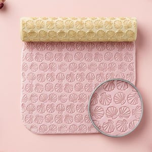 May include: A light pink rolling pin and matching mat with a seashell pattern. The rolling pin is beige and the mat is light pink. A magnifying glass shows the seashell pattern. Dried flowers and lavender sprigs are scattered around the rolling pin and mat.