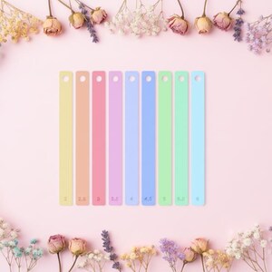 Peut inclure: Huit bracelets en silicone aux couleurs pastel, chacun avec un numéro de mesure différent imprimé dessus. Les bracelets sont disposés sur un fond rose et entourés d'une composition florale de fleurs séchées.