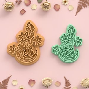 Op de afbeelding: Twee koekjessnijders in de vorm van een hagedis. Eén is bruin, de andere is mintgroen. Beide hebben ingewikkelde bloemen- en bladerpatronen. De snijders zijn omgeven door gedroogde bloemen en bloemblaadjes op een roze achtergrond. Ideaal voor bakken.