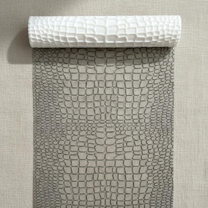 Peut inclure: Un rouleau de papier peint blanc texturé, partiellement déroulé pour révéler un motif gris en forme de peau de crocodile. Le papier peint est présenté sur un fond neutre et texturé.