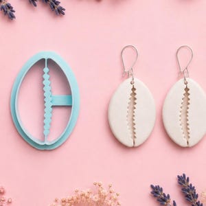 Op de afbeelding: Een lichtblauwe ovale polymeerklei-uitsteker en een paar witte ovale oorbellen met een verticaal zigzagontwerp. De oorbellen hebben zilveren haken. De items liggen op een roze ondergrond met bloemaccenten.