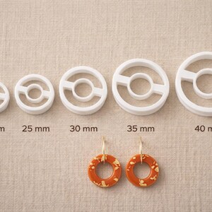 May include: A collection of white, circular polymer clay cutters in various sizes, ranging from 20 mm to 40 mm. Two orange and gold earrings are displayed below the cutters. The background is a neutral beige fabric.