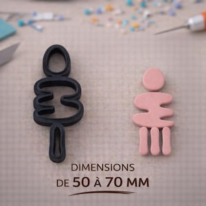 Puede incluir: Un cortador de arcilla polimérica negro y una figura de arcilla rosa. El cortador tiene la forma de una persona estilizada, con dimensiones de 50 a 70 mm. La figura de arcilla rosa está al lado del cortador.
