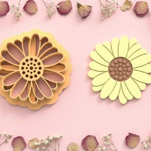 Puede incluir: Dos cortadores de galletas con forma de girasol. Uno es un diseño en capas y abierto en tonos amarillos y melocotón. El otro es un girasol amarillo claro sólido con un centro marrón. El fondo es rosa suave con detalles florales.
