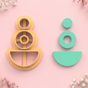 Puede incluir: Dos cortadores de galletas. Uno es de color naranja claro y tiene un diseño apilado con un semicírculo en la parte inferior. El otro es de color verde azulado y tiene un círculo, un anillo y un semicírculo. El fondo es rosa.
