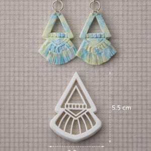 Puede incluir: Pendientes hechos a mano y un cortador a juego. Los pendientes presentan un diseño geométrico con una parte superior triangular y una parte inferior en forma de abanico, en tonos azules, amarillos y blancos. El cortador mide 3,8 cm de ancho y 5,5 cm de alto.
