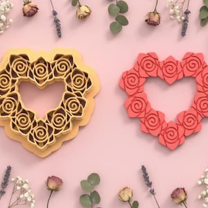 Puede incluir: Dos cortadores de galletas en forma de corazón. Uno es amarillo claro, el otro rojo. Ambos están decorados con diseños de rosas. Los cortadores están sobre una superficie rosa, rodeados de flores secas y vegetación.