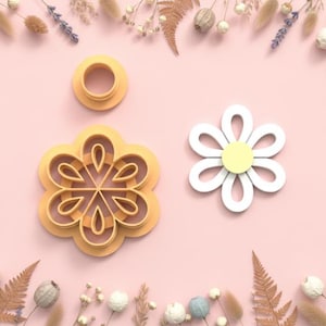 Puede incluir: Un conjunto de tres elementos con forma de margarita. Hay un cortador de galletas de plástico amarillo, un anillo amarillo y una margarita blanca y amarilla. Los artículos están sobre una superficie rosa, rodeados de flores secas.