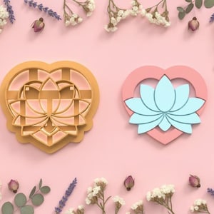 Puede incluir: Dos cortadores de galletas en forma de corazón. Uno es de color marrón claro con un diseño de flor de loto. El otro es un corazón rosa con una flor de loto azul claro en el interior. El fondo es rosa con detalles florales.