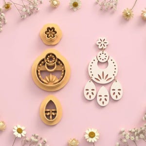 Puede incluir: Un conjunto de cortadores de galletas amarillos con forma de flor y un cortador blanco con forma de pendiente sobre un fondo rosa. Incluye una flor, un círculo floral, un óvalo y un pendiente grande.