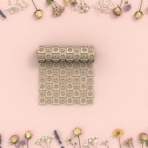 Peut inclure: Un tapis de yoga beige à motifs floraux est déroulé sur une surface rose, entouré de fleurs séchées. Le tapis est composé de petits blocs carrés, chacun avec un motif floral. La partie enroulée du tapis est visible en haut.