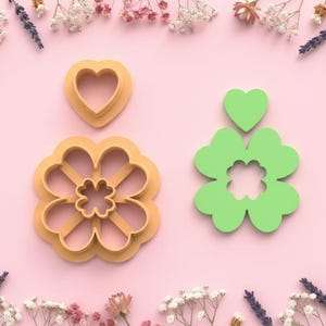 Puede incluir: Tres cortadores de galletas: un corazón, una flor de cuatro pétalos y un trébol de cuatro hojas. Los cortadores de corazón y flor son de color naranja claro, mientras que el trébol es verde. Los artículos están dispuestos sobre una superficie rosa, rodeados de flores.