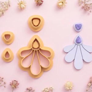 Puede incluir: Una colección de cortadores de arcilla polimérica amarillos y morados claros en varias formas florales, incluyendo una gota, un círculo y un diseño de flor grande. Los cortadores están dispuestos sobre una superficie rosa claro, con flores secas esparcidas.