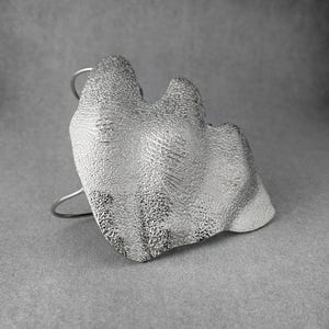 Puede incluir: Un brazalete plateado con un dise&ntilde;o org&aacute;nico y texturizado. El brazalete presenta una forma grande e irregular con una superficie detallada, montada sobre una banda curva simple. La est&eacute;tica general es moderna y escult&oacute;rica.