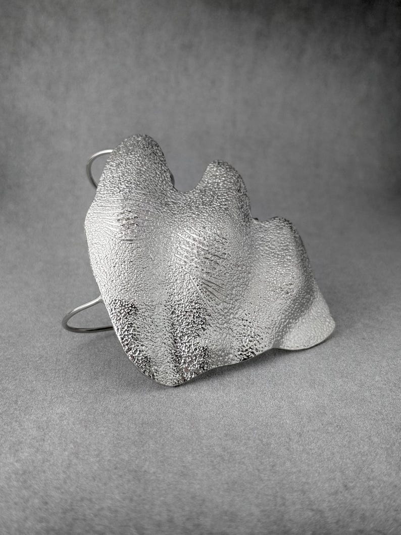 Puede incluir: Un brazalete plateado con un dise&ntilde;o org&aacute;nico y texturizado. El brazalete presenta una forma grande e irregular con una superficie detallada, montada sobre una banda curva simple. La est&eacute;tica general es moderna y escult&oacute;rica.