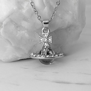 Puede incluir: Collar con colgante plateado con un diseño de cruz y un orbe en forma de planeta, adornado con cristales transparentes. El colgante cuelga de una fina cadena. La joya se muestra sobre un fondo blanco.