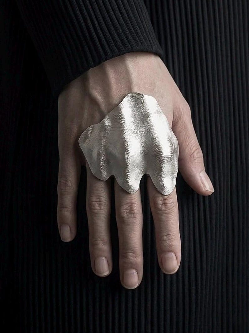 Puede incluir: Un gran anillo plateado con un dise&ntilde;o org&aacute;nico y fluido. El anillo se lleva en una mano, sobre un fondo oscuro y texturizado. La superficie del anillo tiene una textura ondulada, que capta la luz y crea reflejos. Una pieza llamativa.