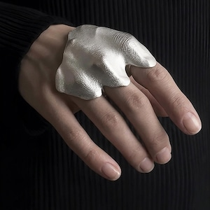 Puede incluir: Un anillo plateado con un dise&ntilde;o org&aacute;nico y texturizado se muestra en una mano. El anillo tiene una forma irregular, que se asemeja a una forma fluida. El fondo es una superficie oscura y texturizada, que contrasta con el brillo met&aacute;lico del anillo.