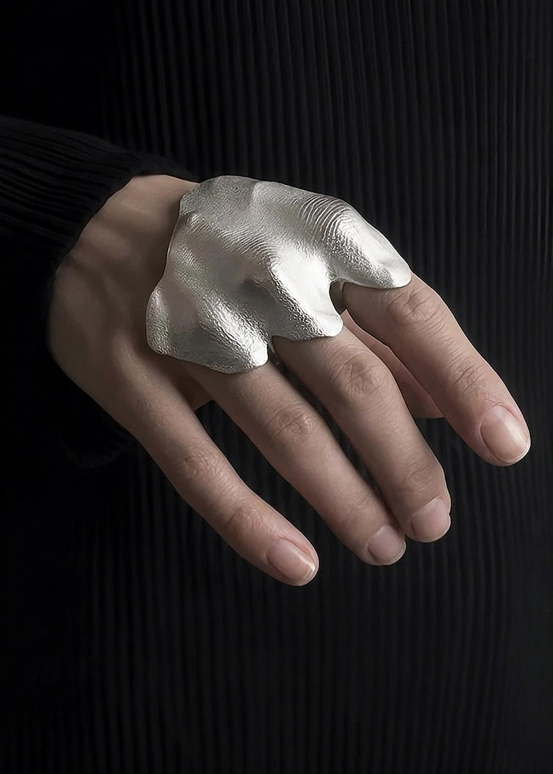 Puede incluir: Un anillo plateado con un dise&ntilde;o org&aacute;nico y texturizado se muestra en una mano. El anillo tiene una forma irregular, que se asemeja a una forma fluida. El fondo es una superficie oscura y texturizada, que contrasta con el brillo met&aacute;lico del anillo.