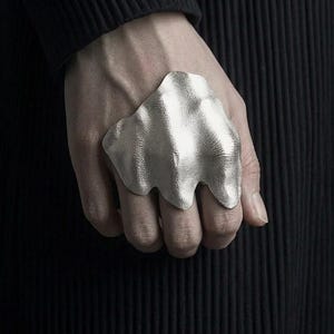 Puede incluir: Un anillo plateado con un dise&ntilde;o org&aacute;nico y fluido. El anillo tiene una superficie texturizada y se lleva en una mano sobre un fondo oscuro. La forma del anillo recuerda a una ola o una forma fundida.