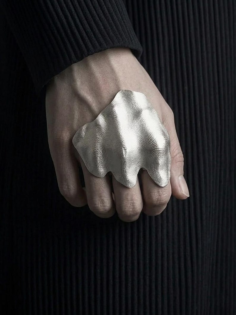 Puede incluir: Un anillo plateado con un dise&ntilde;o org&aacute;nico y fluido. El anillo tiene una superficie texturizada y se lleva en una mano sobre un fondo oscuro. La forma del anillo recuerda a una ola o una forma fundida.