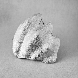 Puede incluir: Un anillo de metal plateado texturizado con un dise&ntilde;o org&aacute;nico en forma de ola. El anillo tiene una forma irregular &uacute;nica y un acabado mate. La joya se encuentra sobre un fondo gris neutro.