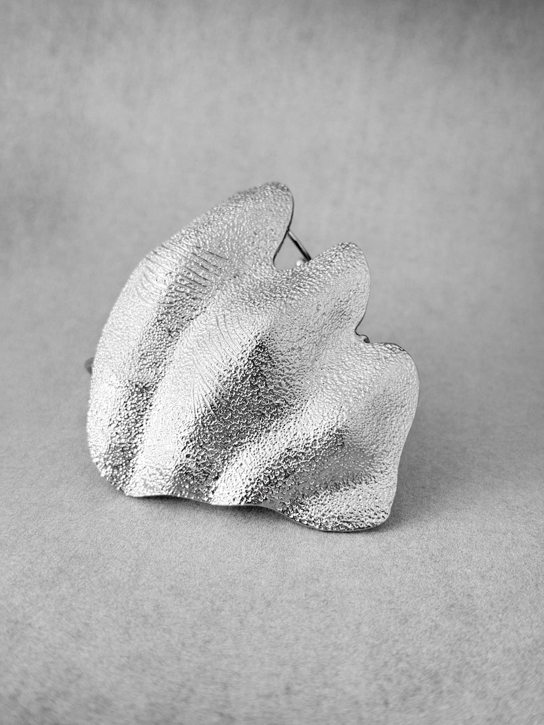 Puede incluir: Un anillo de metal plateado texturizado con un dise&ntilde;o org&aacute;nico en forma de ola. El anillo tiene una forma irregular &uacute;nica y un acabado mate. La joya se encuentra sobre un fondo gris neutro.