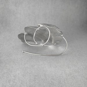 Puede incluir: Un brazalete plateado de dise&ntilde;o abstracto. Presenta una forma texturizada en forma de hoja con tres detalles circulares y una barra curva. El brazalete est&aacute; sobre un fondo gris.