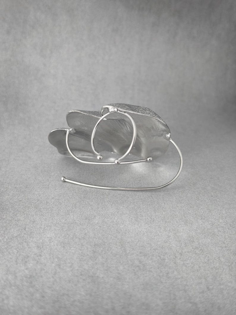 Puede incluir: Un brazalete plateado de dise&ntilde;o abstracto. Presenta una forma texturizada en forma de hoja con tres detalles circulares y una barra curva. El brazalete est&aacute; sobre un fondo gris.