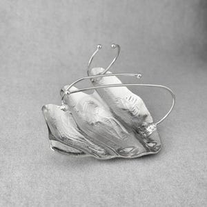 Puede incluir: Broche plateado de dise&ntilde;o abstracto, con forma de hoja. La superficie es texturizada y presenta delicados detalles de alambre, creando una est&eacute;tica moderna y escult&oacute;rica. Sobre un fondo gris neutro.