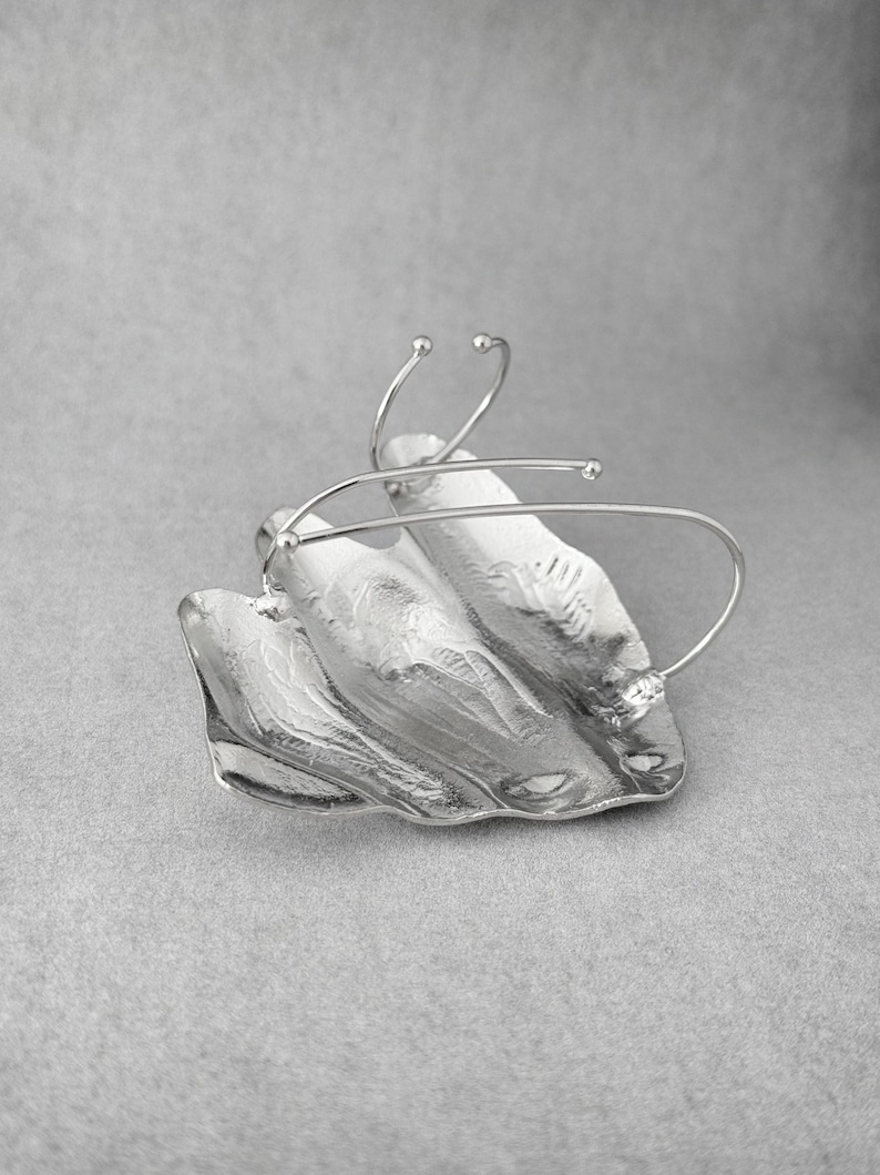 Puede incluir: Broche plateado de dise&ntilde;o abstracto, con forma de hoja. La superficie es texturizada y presenta delicados detalles de alambre, creando una est&eacute;tica moderna y escult&oacute;rica. Sobre un fondo gris neutro.